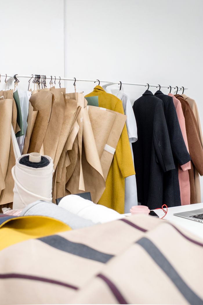 Rack with assorted clothes and sewing patterns on hangers behind various fashion trendy fabrics and textiles placed in modern fashion designer workshop